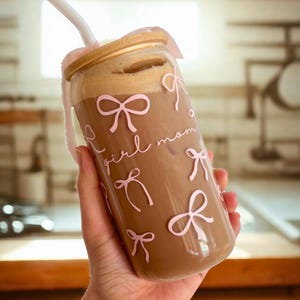 May include: A glass tumbler with a gold lid and a straw. The tumbler is filled with a brown beverage and has a pink and white design with the words "girl mom" and bows.