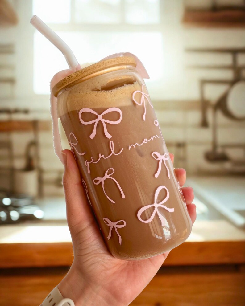 May include: A glass tumbler with a gold lid and a straw. The tumbler is filled with a brown beverage and has a pink and white design with the words "girl mom" and bows.