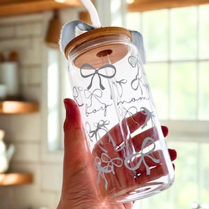 May include: A clear glass reusable water bottle with a bamboo lid and a blue fabric strap. The bottle has a white design of bows and the words "Boy Mom".