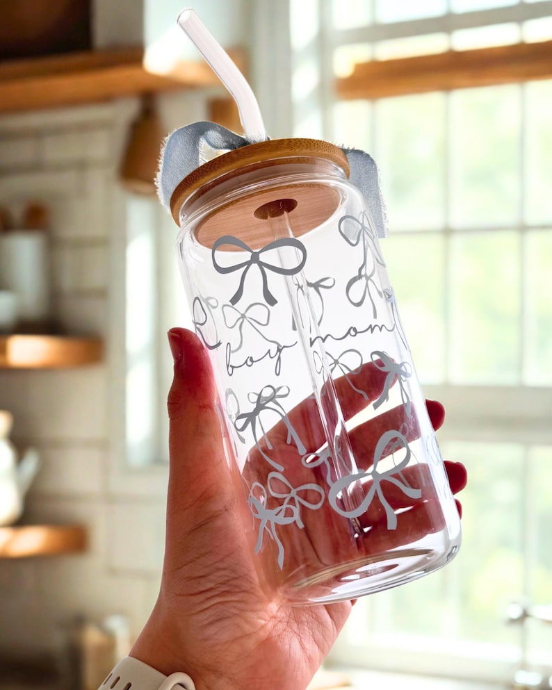 May include: A clear glass reusable water bottle with a bamboo lid and a blue fabric strap. The bottle has a white design of bows and the words "Boy Mom".