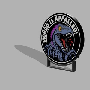 May include: A circular sign featuring a graphic of a blue dinosaur with purple accents, yellow eyes, and an open mouth. The text "MONGO IS APPALLED!" is printed in white capital letters along the top edge of the black circular border frame.