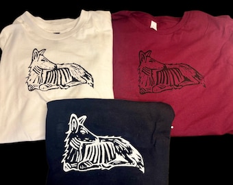 May include: Three short-sleeved t-shirts in white, maroon, and black. Each shirt features a black or white block-print style graphic of a reclining coyote with visible rib cage details, rendered in a stylized, linear artistic fashion.