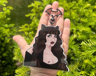 May include: A clear acrylic keychain featuring an illustrated character with long dark wavy hair, cat ears, and a black top. The character has pale skin, red lips with a drop of blood, and sharp fangs. The keychain includes a black bat-shaped clasp.