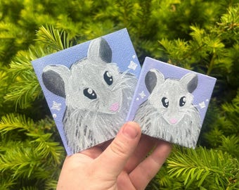 May include: Two small square paintings of a mouse with gray fur, black ears, and a pink nose. The background is a light blue with white star accents. The paintings are held in front of a green, leafy background.