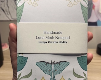 May include: A rectangular notepad featuring a light green luna moth illustration at the bottom and floral accents at the top corners. A white paper band wraps around the center with the text: Handmade Luna Moth Notepad Creepy Crawlin Oddity.