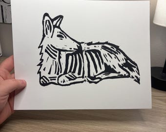 May include: A black and white linocut print on white paper featuring a stylized illustration of a canine lying down. The artwork depicts the animal with a visible ribcage and skeletal structure, rendered in bold, high-contrast ink lines.
