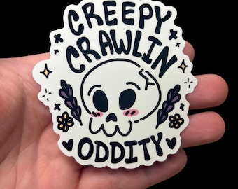 May include: A die-cut sticker featuring a cartoon skull with pink cheeks, surrounded by small purple floral sprigs and yellow stars. The text reads CREEPY CRAWLIN ODDITY in bold black lettering. The sticker has a white background and scalloped edges.