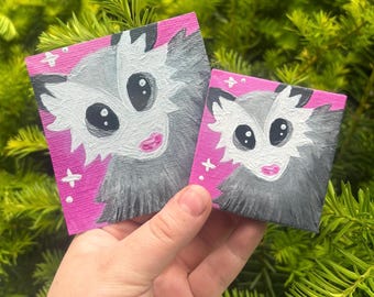 May include: Two small square paintings featuring opossum portraits. The opossums have large black eyes, pink lips, and gray fur, set against a bright pink background. White accents and small crosses add detail. The paintings are held against a backdrop of green foliage.