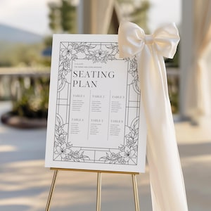 May include: A white wedding seating plan on a gold easel, adorned with floral illustrations and the text "SEATING PLAN." A cream-colored bow and fabric drape from the top. The sign lists table numbers and guest names.
