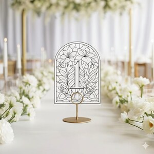 May include: A table number card with the number 1, featuring a floral design. The card is held by a gold-colored metal stand. The background shows a white table setting with flowers.