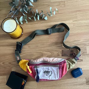 May include: A multi-colored fanny pack with a green strap and buckle. The pack has a white patch with the word "Vintage" and other text. A black wallet and blue earbud case are also visible.
