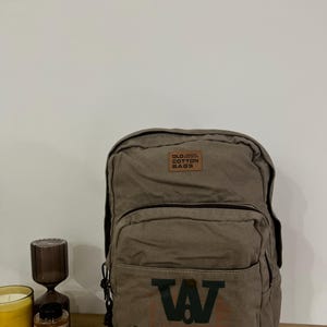 May include: An olive green canvas backpack with a front pocket displaying a dark green "W" and the words "OLD COTTON VINTAGE" in orange. The top has a small leather patch with the words "OLD COTTON BAGS".