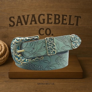 Puede incluir: Un cinturón de cuero color verde azulado con detalles florales en relieve. El cinturón presenta hebillas doradas y verde azulado ornamentadas y una trabilla a juego. El texto "SAVAGEBELT CO." está encima del cinturón y "© 2022 SAVAGEBELT CO." debajo.