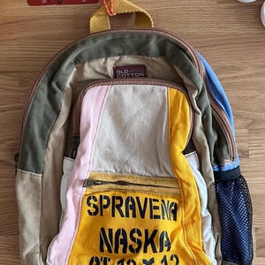 Puede incluir: Una mochila multicolor con una variedad de telas y texturas. El bolsillo delantero presenta un panel amarillo con texto negro que dice "SPRAVENA NASKA AT 12 X 12". La bolsa tiene un bolsillo lateral de malla y un asa superior.