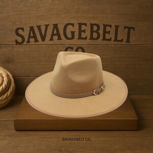 May include: A tan fedora hat with a light brown leather band and buckle. The hat has a wide brim and a structured crown. The text "SAVAGEBELT CO." is visible in the background. The hat is displayed on a wooden surface.