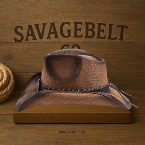 May include: A brown cowboy hat with a distressed finish and a black leather band. The hat is sitting on a wooden shelf with the text "SAVAGEBELT CO." printed below.