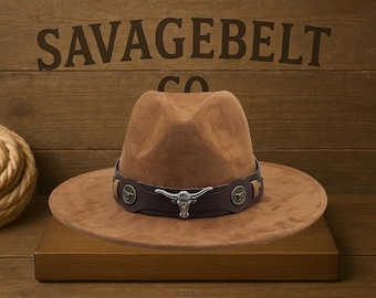 Pode incluir: Um chapéu fedora de camurça marrom com uma faixa de couro marrom e uma fivela de prata com cabeça de touro. O chapéu tem o texto "SAVAGEBELT CO." na faixa.