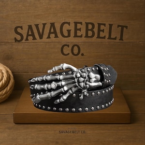 Puede incluir: Cinturón de cuero negro con hebilla de mano de esqueleto plateada y tachuelas. El cinturón se muestra sobre una superficie de madera con el texto "SAVAGEBELT CO." y "©2022 SAVAGEBELT CO."