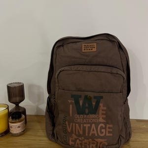 Może przedstawiać: Brązowy plecak z płótna z przednią kieszenią i napisem "OLD FABRIC CREATIONS VINTAGE FABRIC". Plecak ma małą brązową skórzaną naszywkę z napisem "OLD COTTON BAGS".