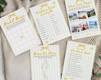 May include: A collection of bridal shower game cards with gold text and accents. The cards include games like "Bridal Bingo," "Wedding Word Scramble," and "Guess the Dress." The cards are arranged on a neutral-toned fabric.