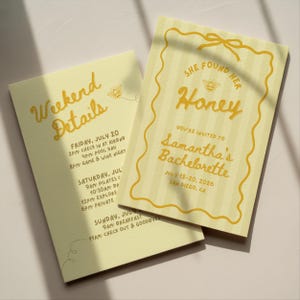 May include: Two pale yellow rectangular invitation cards with vertical stripes and dark yellow text. One card reads 'She Found Her Honey' with a bee graphic, and the other is titled 'Weekend Details' listing a schedule for July 20-22, 2026.