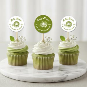 May include: Three matcha cupcakes with white frosting and green buttercream swirls, topped with decorative Baby Green toppers. Each cupcake has a sprig of baby's breath and a mint leaf. The toppers read "Baby Green" and "We love you so matcha!"