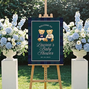 May include: A wooden easel displays a baby shower sign with a navy blue and green plaid border. The sign reads "Welcome to Lauren's Baby Shower" with two teddy bears wearing sweaters with American flags. The date 09.18.2025 is at the bottom.