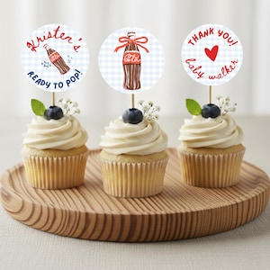 May include: Three cupcakes with white icing, blueberries, and decorative toppers. The toppers feature Coca-Cola bottle illustrations and text that reads "Kristen's Ready to Pop!", "Coca-Cola", and "Thank You! Baby Walker". The cupcakes are on a wooden tray.