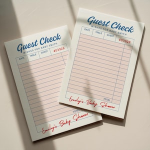 May include: Two guest check pads with the text "Guest Check" in blue script. The pads have pink horizontal lines and the text "Wishes for Baby Smith". The bottom of the pads read "Emily's Baby Shower".