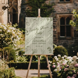 May include: A wooden easel displays a baby shower sign with the text "Locally Grown Baby!" and "Welcome to Samantha's Baby Shower 05.14.26." The sign has a light green background with vertical stripes and illustrations of vegetables and a bow.