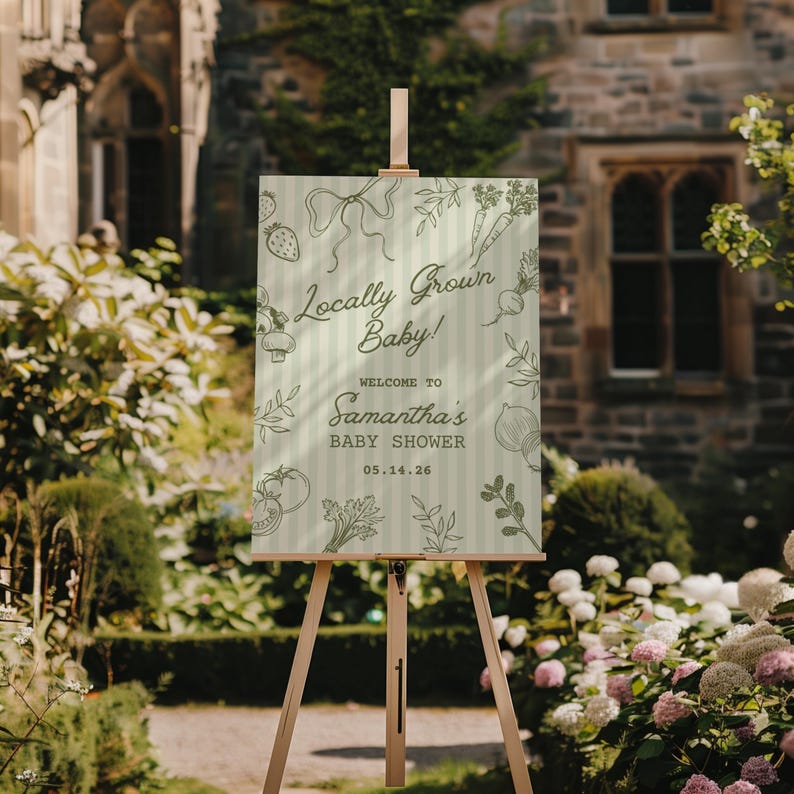 May include: A wooden easel displays a baby shower sign with the text "Locally Grown Baby!" and "Welcome to Samantha's Baby Shower 05.14.26." The sign has a light green background with vertical stripes and illustrations of vegetables and a bow.