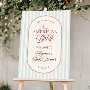 May include: A baby shower welcome sign on a wooden easel. The sign has a light blue and white striped background with a red oval frame. Text includes "Born in the U.S.A. All American Baby" and "Welcome to Meghan's Baby Shower 07.15.26."