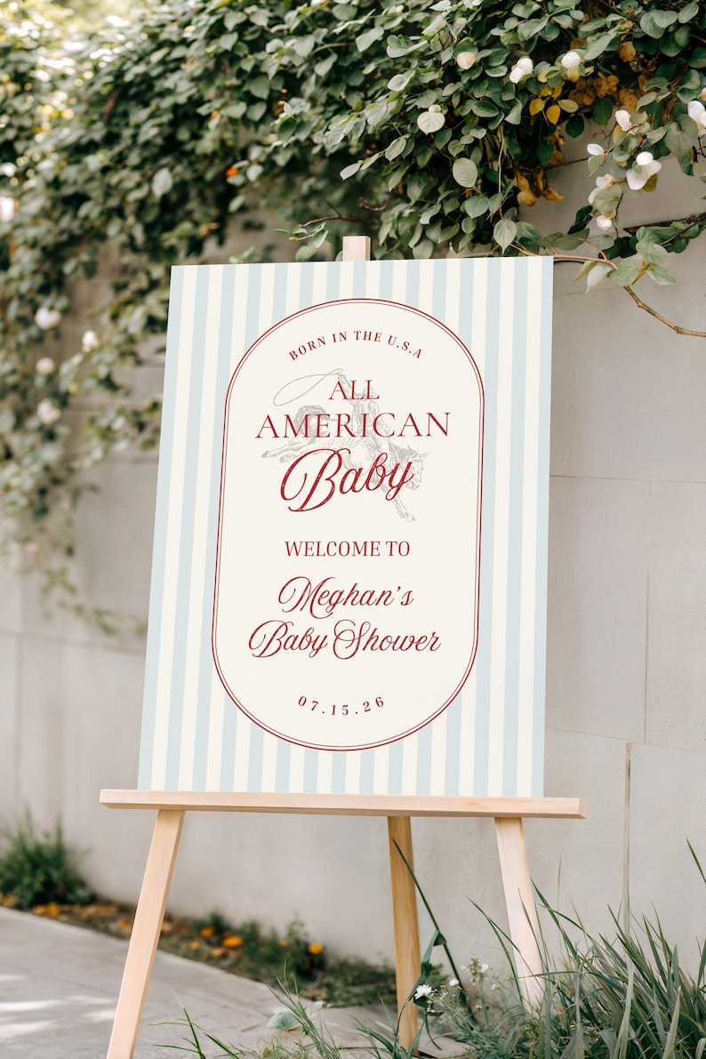 May include: A baby shower welcome sign on a wooden easel. The sign has a light blue and white striped background with a red oval frame. Text includes "Born in the U.S.A. All American Baby" and "Welcome to Meghan's Baby Shower 07.15.26."