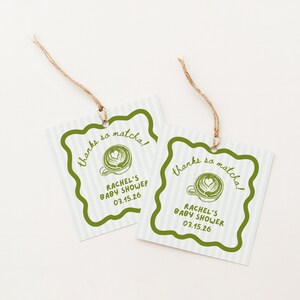 May include: Two square gift tags with a green scalloped border and a light blue striped background. The tags read "thanks so matcha!" and "RACHEL'S BABY SHOWER 03.15.26" with a matcha latte graphic. Each tag has a twine string.