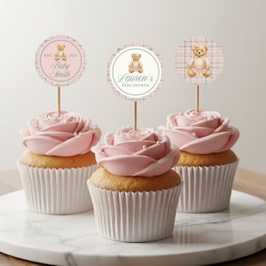 May include: Three cupcakes with pink rose-shaped frosting and pearl sprinkles. Each cupcake has a circular topper with a teddy bear illustration and text that reads "Baby Smith", "Lauren's Baby Shower", and a teddy bear in a dress.