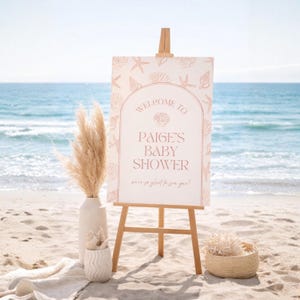 May include: A baby shower welcome sign on a wooden easel, with the text "Welcome to Paige's Baby Shower". The sign is decorated with seashell illustrations. The scene is set on a sandy beach with the ocean in the background.