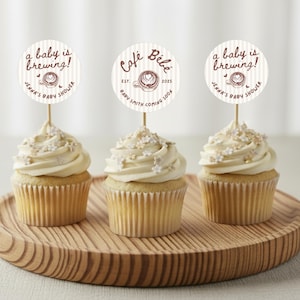 May include: Three cupcakes with white frosting and decorative sprinkles sit on a wooden tray. Each cupcake has a circular topper with text that reads "a baby is brewing!" and "Jenna's Baby Shower."