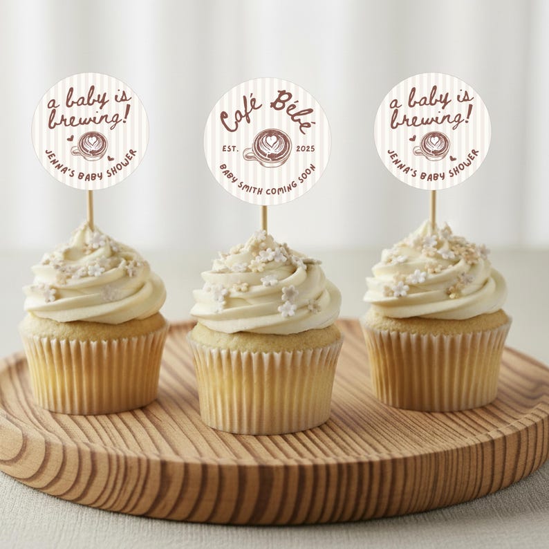 May include: Three cupcakes with white frosting and decorative sprinkles sit on a wooden tray. Each cupcake has a circular topper with text that reads "a baby is brewing!" and "Jenna's Baby Shower."