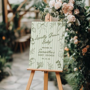 May include: A wooden easel displays a baby shower sign with the text "Locally Grown Baby!" and "Welcome to Samantha's Baby Shower" in green script. The sign has a light green striped background and illustrations of vegetables.
