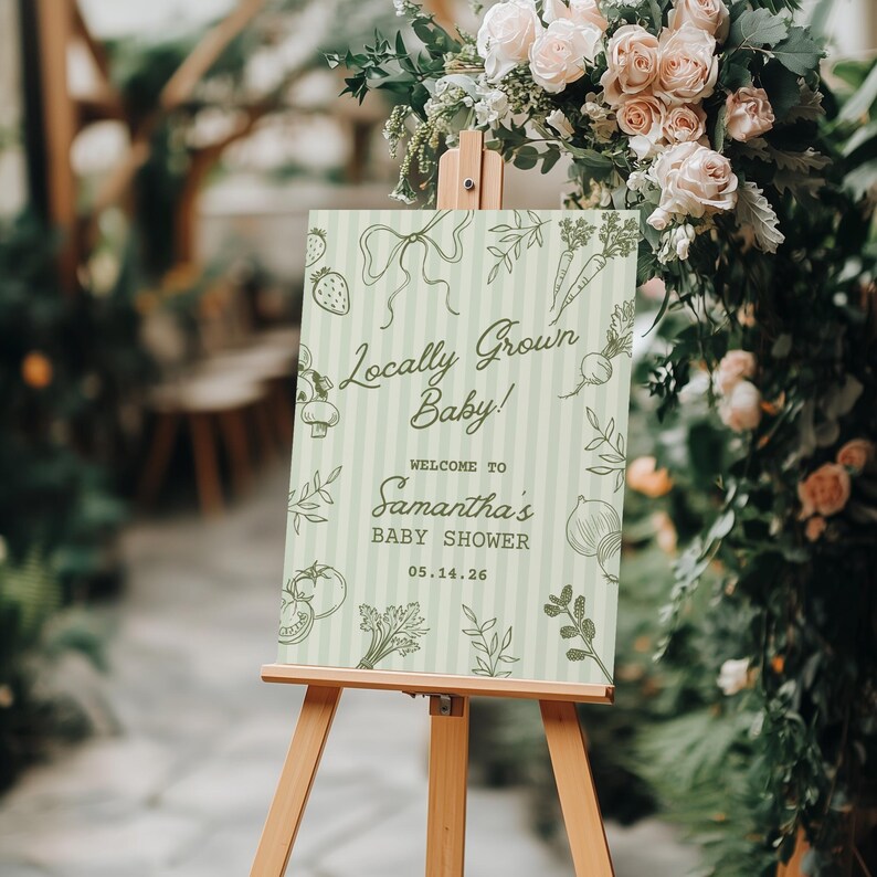 May include: A wooden easel displays a baby shower sign with the text "Locally Grown Baby!" and "Welcome to Samantha's Baby Shower" in green script. The sign has a light green striped background and illustrations of vegetables.