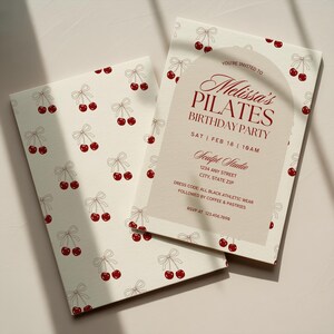 May include: Two cream-colored invitations with a cherry pattern. One invitation is open, revealing text that reads "Melissa's Pilates Birthday Party" with event details. The cherry design includes red cherries and light brown bows.