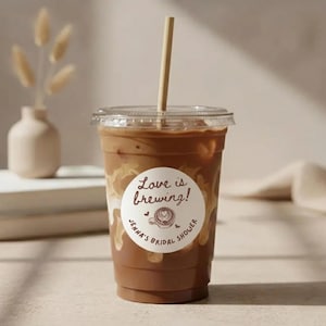 May include: A clear plastic cup filled with iced coffee, a straw, and a lid. A white sticker on the cup reads "Love is Brewing!" with "Jenna's Bridal Shower" written around a coffee cup graphic. The coffee has a creamy, layered appearance.