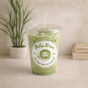May include: A clear plastic cup filled with a green matcha drink, topped with a clear lid and a straw. The cup has a white sticker with the words "Baby Green" and "We love you so matcha!" The drink is next to a small potted plant.