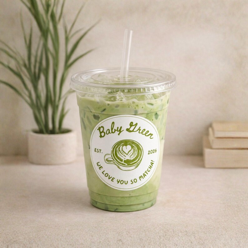 May include: A clear plastic cup filled with a green matcha drink, topped with a clear lid and a straw. The cup has a white sticker with the words "Baby Green" and "We love you so matcha!" The drink is next to a small potted plant.