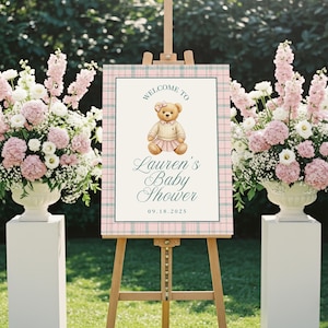 May include: A baby shower sign on a wooden easel, featuring a teddy bear illustration. The sign has a pink and green plaid border and reads "Welcome to Lauren's Baby Shower" with the date 09.18.2025. Floral arrangements in white urns flank the sign.