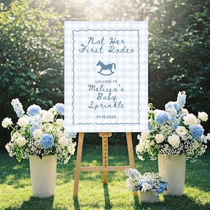 May include: A baby sprinkle welcome sign on a wooden easel, with the text "Not Her First Rodeo" and "Welcome to Melissa's Baby Sprinkle 01.18.2026". The sign has a blue gingham border and a rocking horse graphic. White and blue floral arrangements flank the sign.