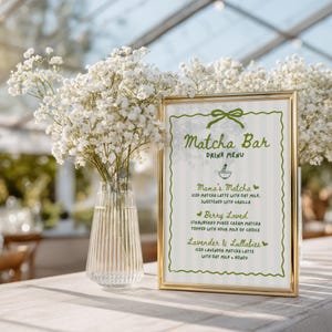 May include: A gold-framed "Matcha Bar Drink Menu" sign with a green bow design. The menu lists "Mama's Matcha," "Berry Loved," and "Lavender & Lullabies" drinks. A clear glass vase with white flowers is to the left.
