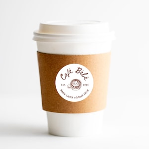May include: A white paper coffee cup with a brown cardboard sleeve. A round sticker on the cup reads "Caf&eacute; B&eacute;b&eacute;" with a coffee cup design and the text "Baby Smith Coming Soon". The cup has a white lid.