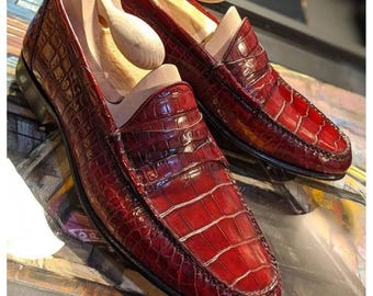 Könnte beinhalten: Ein Paar burgunderfarbene Lederschuhe mit Kroko-Muster. Die Loafer haben eine spitze Zehenpartie und einen niedrigen Absatz.