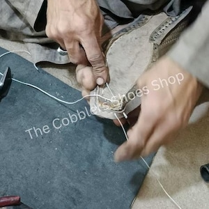 May include: A cobbler's hands working on a shoe, using a hammer and white thread. The image shows the process of shoe repair or construction. The text "The Cobblers Shoes Shop" is visible on the surface.